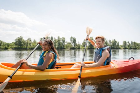 feliz y encantadora mujer afroamericana con hombre joven y pelirrojo sosteniendo paletas y navegando en kayak deportivo en el lago escénico en fin de semana de verano activo