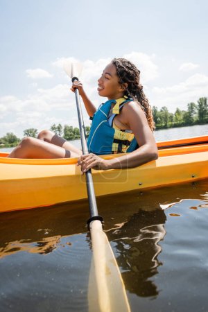 Foto de Vista lateral de morena y atractiva mujer afroamericana en chaleco salvavidas sosteniendo remo y navegando en kayak deportivo durante la recreación acuática en verano - Imagen libre de derechos