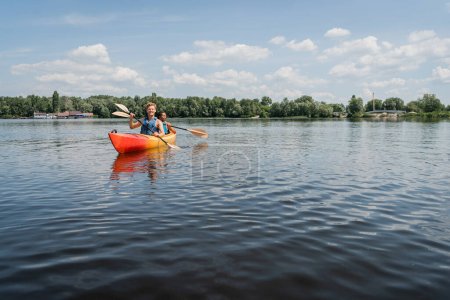 Aktives multiethnisches Paar in Sicherheitswesten segelt im sportlichen Kajak auf malerischem See mit grünem Ufer bei blauem und bewölktem Himmel während der Wassererholung am Sommerwochenende