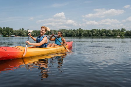 happy and sportive interracial couple in life vests spending vacation on picturesque lake and sailing in kayak with paddles under blue sky with clouds