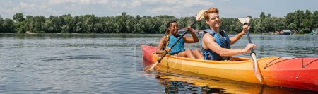 Photo for Joyful redhead man and pretty african american woman in life vests paddling in sportive kayak during summer recreation on river with green trees on bank, banner - Royalty Free Image