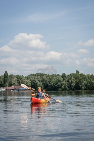 Foto de Deportiva pareja multiétnica en chalecos salvavidas remando en kayak en el río con verde pintoresco banco bajo el cielo azul nublado durante el fin de semana de recreación en verano - Imagen libre de derechos