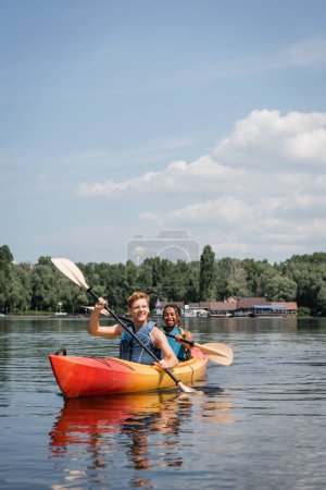 glückliches und aktives multiethnisches Paar in Schwimmwesten, das im sportlichen Kajak mit Paddeln auf dem Fluss mit grünem Ufer unter blauem Himmel mit Wolken im Sommer segelt