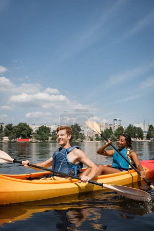 positive and active multiethnic couple in life vests spending recreation weekend by sailing in sportive kayak on city river under blue sky with white clouds