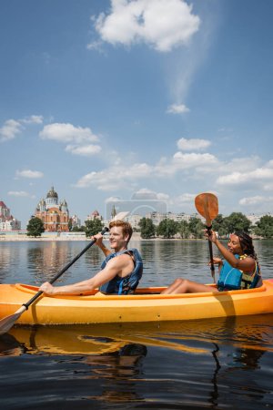 Photo for Carefree redhead man and pretty african american woman in life vests sailing in sportive kayak along riverside with cityscape and green trees under blue and cloudy sky - Royalty Free Image