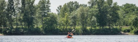 Photo for View from afar of interracial active couple in life vests sailing in sportive kayak with paddles near riverside with green trees on summer day, banner - Royalty Free Image
