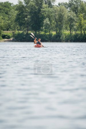 vista desde lejos de la pareja multiétnica deportiva en chalecos salvavidas sosteniendo paletas mientras navega en kayak en el lago con la costa pintoresca verde en el fin de semana de verano