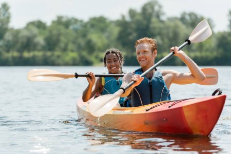hombre pelirrojo activo y encantadora mujer afroamericana en chalecos salvavidas pasar tiempo en el río mientras navega en kayak deportivo en el pintoresco lago sobre fondo borroso en verano