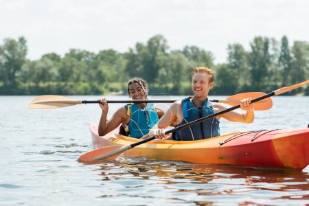 young redhead man in life vest paddling in kayak on summer day near cheerful and pretty african american woman on river with green bank on blurred background