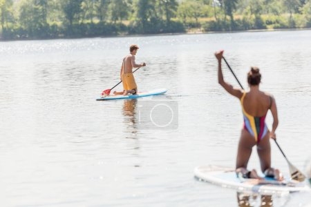 Foto de Joven hombre deportivo navegando en sup board en el pintoresco lago cerca de la mujer afroamericana en traje de baño colorido paddleboarding en primer plano borroso en verano - Imagen libre de derechos