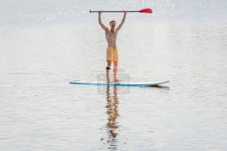 volle Länge eines überglücklichen rothaarigen Mannes in gelben Badehosen mit Paddel in erhobenen Händen, während er am Sommerwochenende auf einem Surfbrett auf einem See mit ruhigem Wasser steht