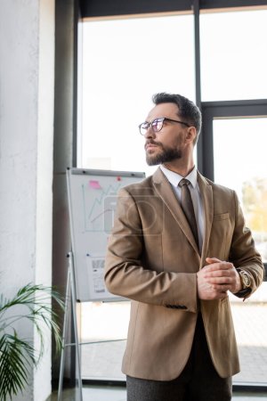 thoughtful bearded corporate manager in trendy formal wear and eyeglasses standing near blurred flip chart with business infographics and looking away in office 