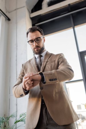 Tiefansicht eines stilvollen bärtigen Unternehmensmanagers in trendigem beigen Blazer, Krawatte und Brille, der auf eine Armbanduhr blickt, während er auf ein Meeting im Büro wartet 