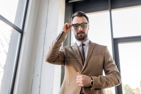 Tiefansicht des ehrgeizigen und erfolgreichen bärtigen Geschäftsmannes in beige stylischem Blazer und Krawatte, der Brille anpasst und im modernen Büro in die Kamera blickt 