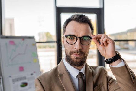 bearded successful businessman in beige blazer and tie adjusting stylish eyeglasses and looking at camera near flip chart with graphs on blurred background 