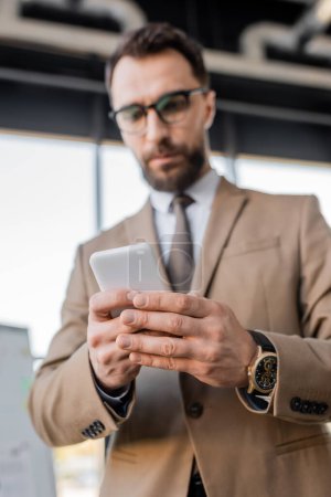 Low-Winkelbild eines seriösen verschwommenen Geschäftsmannes in Luxus-Armbanduhr, beige stylischem Blazer, Brille und Krawatte mit Handy und Denken im Büro