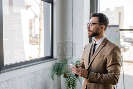 ernster bärtiger Geschäftsmann in beigem Blazer und Brille, Smartphone in der Hand, während er durchs Fenster schaut und in der Nähe von Flipchart und Topfpflanze im Büro denkt