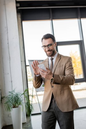 alegre gerente barbudo en elegante traje de negocios y anteojos saludando la mano durante la videollamada en el teléfono celular cerca de rotafolio y maceta planta en la oficina moderna