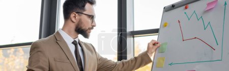 thoughtful and ambitious businessman in beige stylish blazer, tie and eyeglasses attaching sticky note on flip chart with infographics in office, banner