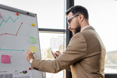 Photo for Side view of accomplished businessman in beige suit and eyeglasses holding mobile phone and drawing on flip chart while making productivity analysis in office - Royalty Free Image
