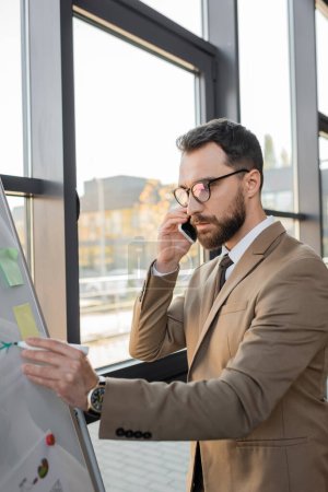 serious bearded entrepreneur in stylish eyeglasses and beige blazer talking on mobile phone and drawing with marker on flip chart while doing business analytics in office