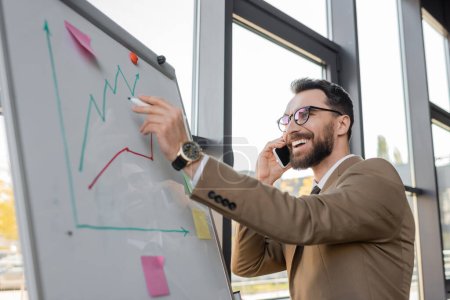 happy and satisfied businessman in eyeglasses and beige suit talking on mobile phone while standing with marker near flip chart and looking at increasing graphs in office