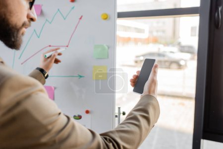 cropped view of bearded businessman in beige blazer holding mobile phone with blank screen and marker while drawing graphs on flip chart with sticky notes in office