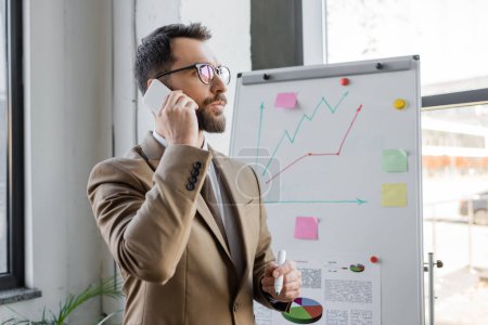 thoughtful manager in stylish business attire and eyeglasses standing with marker near analytics and graphs on flip chart and talking on mobile phone in office