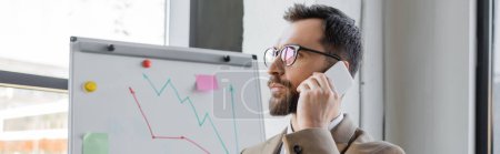 portrait of thoughtful bearded businessman in stylish eyeglasses talking on mobile phone and looking away near flip chart with analytics in office, banner