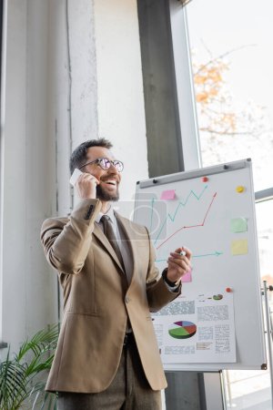 joyful bearded manager in beige blazer, tie and eyeglasses standing with marker and talking on mobile phone near flip chart with business analytics and graphs in office