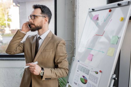 thoughtful bearded businessman in beige blazer and eyeglasses holding marker and looking away while talking on smartphone near market research on flip chart in office