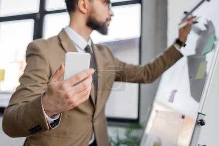 Geschäftsmann im eleganten beigen Blazer, Smartphone in der Hand und mit Filzstift auf Flipchart schreiben, während er im Büro Produktivitätsanalysen durchführt, verschwommener Hintergrund