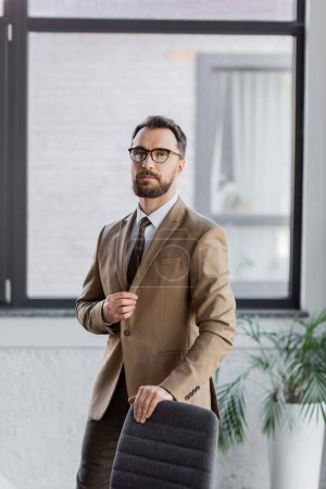 homme d'affaires confiant et à la mode dans les lunettes, blazer beige et cravate debout près de la chaise et regardant la caméra dans le bureau contemporain sur fond flou