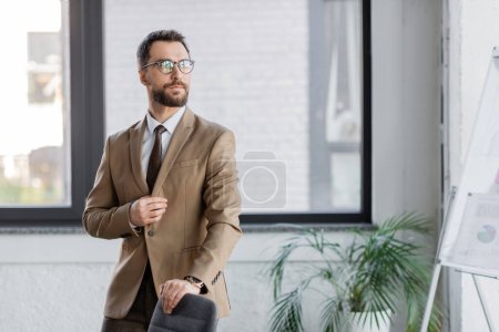 Selbstbewusster und bärtiger Geschäftsmann in beigem Blazer, Krawatte und Brille neben Bürostuhl, verschwommenem Flipchart und Topfpflanze und wegschauendem Blick