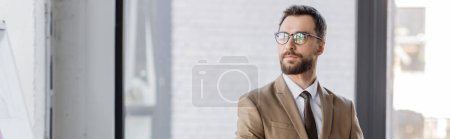 homme d'affaires barbu accompli et confiant en blazer beige, cravate et lunettes élégantes détournant les yeux tout en se tenant debout dans le bureau contemporain sur fond flou, bannière