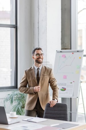 charismatic and satisfied businessman in stylish formal wear and eyeglasses smiling in front of flip chart with graphs near work desk with laptop, coffee cup, notebook and documents in office