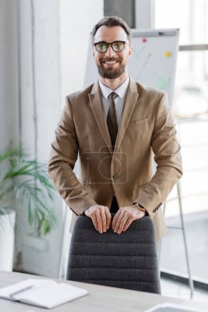 fröhlicher gutaussehender Geschäftsmann in beigem Blazer, Krawatte und Brille, der neben Bürostuhl steht und vor Flipchart auf verschwommenem Hintergrund in die Kamera lächelt