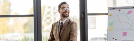 successful bearded businessman in stylish eyeglasses, beige blazer and tie looking away and smiling near flip chart with graphs and sticky notes in office with large windows, banner