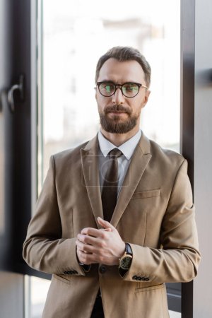 ambicioso hombre de negocios barbudo en blazer beige, gafas con estilo y reloj de pulsera de lujo mirando a la cámara mientras está de pie con las manos cerradas cerca de la ventana en la oficina