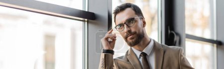 hombre de negocios barbudo en gafas de moda, chaqueta beige y corbata agarrado de la mano cerca de la cara, mirando hacia otro lado y pensando cerca de las ventanas en la oficina moderna, pancarta