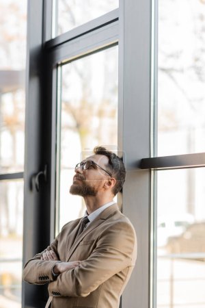 fashionable and dreamy businessman in beige blazer and stylish eyeglasses standing with folded arms near large windows and looking up in modern office