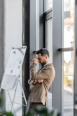 Photo for Side view of exhausted manager in trendy business attire holding eyeglasses and standing with closed eyes near large windows and flip chart with sticky notes in office - Royalty Free Image