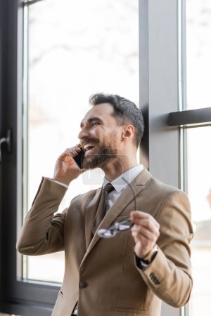 überglücklicher und erfolgreicher Unternehmer im trendigen beigen Blazer und Krawatte im Gespräch mit dem Handy und mit verschwommener Brille vor dem Fenster im modernen Büro