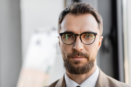portrait of brunette and bearded successful businessman in eyeglasses and stylish formal wear looking at camera in modern office on blurred background