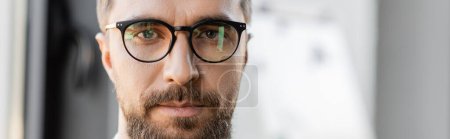 Photo for Portrait of good looking and ambitious businessman with beard and mustache wearing fashionable eyeglasses and looking at camera in blurred office, banner - Royalty Free Image