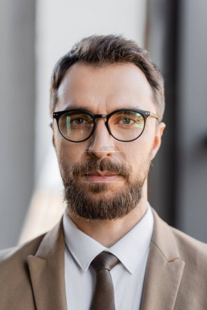 Porträt eines selbstbewussten und charismatischen Geschäftsmannes in stilvoller formaler Kleidung wie beigem Blazer, Krawatte und Brille, der auf verschwommenem Hintergrund in die Kamera im Büro blickt