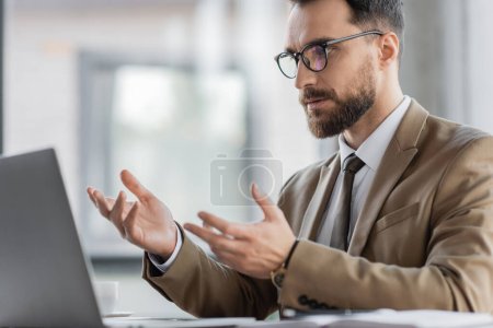 Foto de Hombre de negocios ambicioso y barbudo en blazer elegante beige, corbata y gafas gestos durante la videoconferencia en el ordenador portátil mientras está sentado en el escritorio en la oficina - Imagen libre de derechos