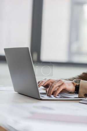 Teilansicht des stilvollen und erfolgreichen Unternehmensmanagers in beige-formaler Kleidung und luxuriöser Armbanduhr, die auf Laptop auf Schreibtisch im Büro tippt, verschwommener Hintergrund