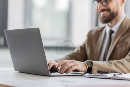 Photo for Cropped view of smiling and successful bearded businessman in beige blazer and stylish eyeglasses chatting on laptop near notebook and pen on office desk - Royalty Free Image