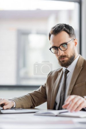 concentrated corporate manager with beard and eyeglasses, wearing trendy blazer and tie, looking at blurred notebook and working on computer in office